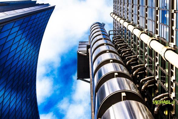 A striking low-angle view of two contrasting London skyscrapers—the curved glass facade of the Leadenhall Building (Cheesegrater) on the left and the industrial, pipe-covered exterior of the Lloyd's Building on the right. The deep blue sky enhances the futuristic and architectural contrast.