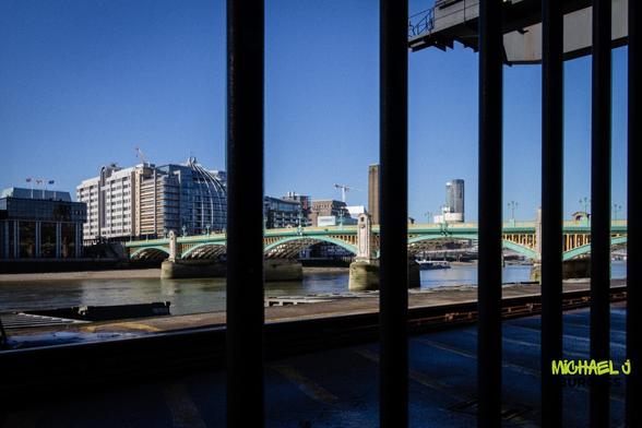 This is a scenic view of Southwark Bridge in London, framed by the bars of a railing. The bridge's green and gold structure contrasts against the blue sky, with modern and historical buildings in the background. The perspective gives a sense of depth and an intriguing composition.
