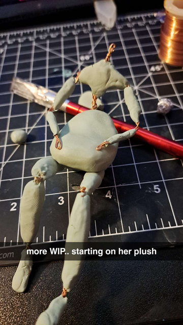 A photo of an armature loosely covered in polymer clay, with a lump of clay in its lap implied to be a plushie. Text on the photo reads "More WIP... starting on her plush"
