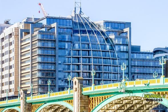 This is a close-up view of Southwark Bridge in London. The vibrant green and gold detailing contrasts with the modern glass-fronted apartments behind it. The curved architectural design, highlighted by a metal framework, reflects the city's historic and contemporary styles.