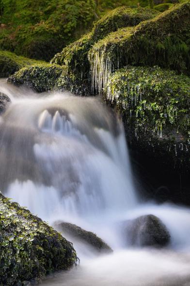 Zu sehen ist eine Wasserfallkaskade mit bemoosten Steinen und Eiszapfen