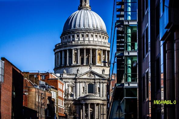 This is a striking view of St. Paul's Cathedral in London, framed by a mix of modern glass buildings and historic brick structures. The composition contrasts the cathedral's timeless grandeur against contemporary architecture's sleek, reflective surfaces.