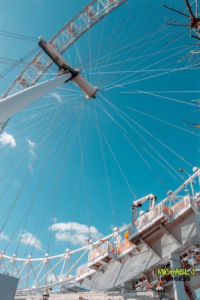 A striking low-angle view of the London Eye, highlighting its intricate cable system against a vibrant blue sky. The structure's support beams and boarding platform are also visible, adding depth and perspective to this iconic landmark.