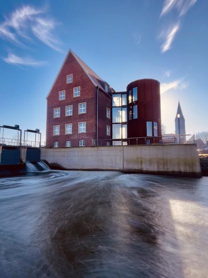 Das Bild zeigt ein Backsteingebäude mit einem modernen Anbau, das an einem Fluss liegt. Das ältere Gebäude ist rotbraun und hat Giebeldach. Der Anbau ist ein runder, dunklerer Bau mit großen Fenstern. Vor dem Gebäude befindet sich eine Betonmauer, hinter der sich ein Wasserlauf befindet, der in Bewegung erscheint. Im Hintergrund ist ein Kirchturm sichtbar. Der Himmel ist klar und blau mit ein paar Wolken. Das Bild vermittelt einen ruhigen und etwas industriellen Eindruck.

Bereitgestellt von @altbot, generiert mit Gemini