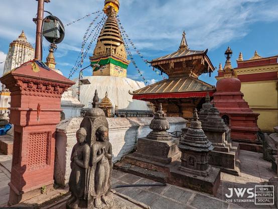 The image depicts the Swayambhunath temple complex, also known as the Monkey Temple, located in Kathmandu, Nepal. The complex is a UNESCO World Heritage Site and one of the most sacred Buddhist sites in Nepal.

In the foreground, there are several stone statues of Buddha and other deities, as well as intricate carvings on the walls and roofs of the temples. The main temple, which is dedicated to the deity Swayambhu, or "Self-Created," is surrounded by smaller shrines and stupas. The complex also features a large stupa at its center, which is adorned with colorful prayer flags and offers stunning views of the city.

The image also shows several monkeys roaming around the temple grounds, adding to the unique atmosphere of this sacred site. Overall, the Swayambhunath temple complex is a must-visit destination for anyone interested in exploring Nepal's rich cultural heritage and spiritual traditions.