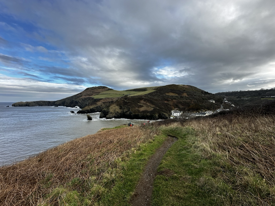 Coastal landscape with a path leading to a small bay surrounded by hills. A few people are visible near the water. Cloudy sky above.