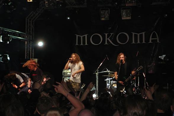 Mokoma esiintymässä Tuskan teltassa olevalla lavalla 2004. Bändin takana seinälakana jossa lukee Mokoma. Kuvan etualalla yleisössä muutamia käsiä ylhäällä.