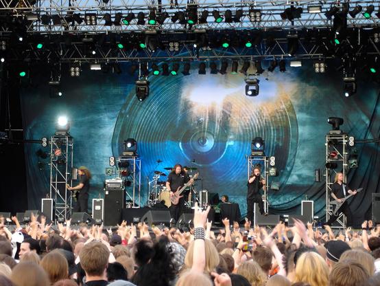 In Flames esiintymässä Tuskan päälavalla 2004. Bändin takana lavan peittävä lakana jossa Soundtrack to Your Escape levyn kannen kuva. Lavan edessä olevassa yleisössä lähes kaikilla on käsi yläällä osoittamassa suosiota bändille.