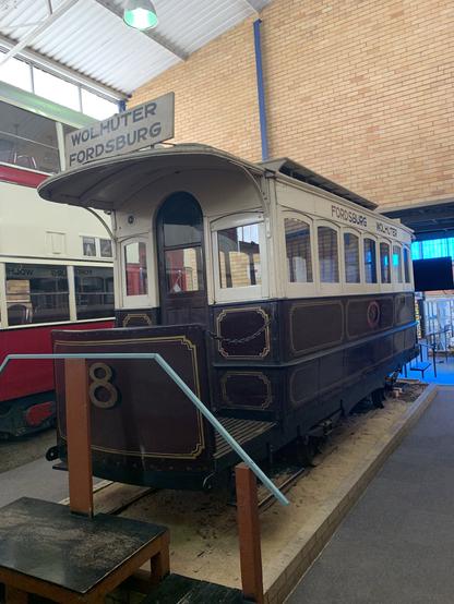 A vintage tram displayed indoors, featuring the signs "Wolhuter" and "Fordsburg." The tram has a wooden exterior with intricate details and is positioned on a raised platform, surrounded by other historical transport exhibits.