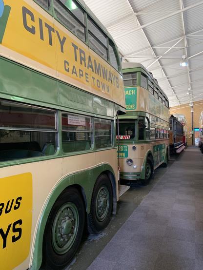 A row of vintage buses exhibits the "City Tramways" branding, prominently displaying "Cape Town" on their sides. The buses are parked inside a spacious facility with a high ceiling and well-lit interior.