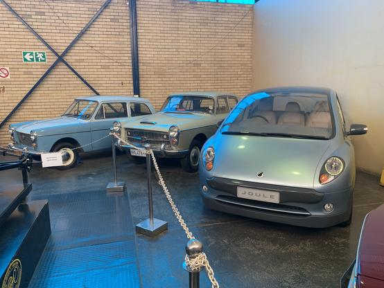 The image features three vintage cars displayed in a museum setting. On the left are two light blue classic cars, while on the right is a modern gray vehicle labeled "Joule." The background includes a brick wall and safety exit signs.