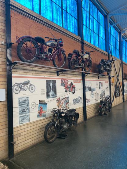 An interior display featuring several vintage motorcycles. Some bikes are mounted on the wall, while others are positioned on the floor against a backdrop of historical images and advertisements related to motorbikes. The space has large windows allowing natural light to filter through.