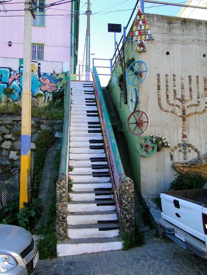 A vibrant set of stairs in Valparaíso, Chile, painted to resemble piano keys, creating a striking musical illusion. The stairs are flanked by stone-lined railings and colorful murals, with one wall adorned with decorative elements like wagon wheels, small figurines arranged in a triangular formation, and a menorah-like design made from stones. Overhead wires crisscross the sky, adding to the urban charm of this artistic location. Parked cars and nearby buildings painted in pastel colors contribute to the lively atmosphere of this cultural hub known for its street art.