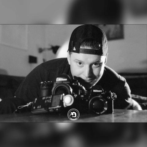 black and white photo of an out-of-focus man wearing a trucker cap with two cameras and a light meter in front of him.