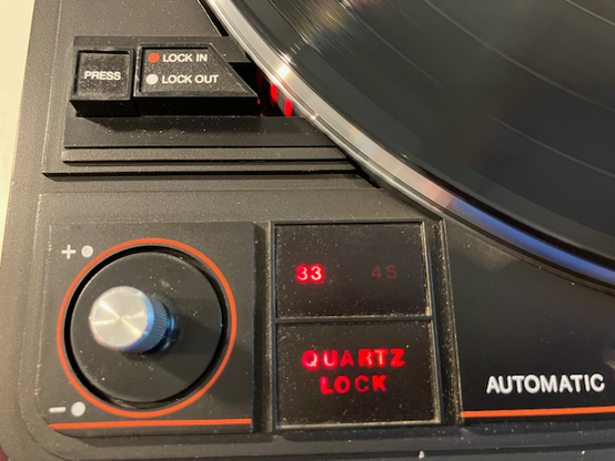 This image shows a close-up of the control panel on the ADC turntable. The features visible include:

- A "PRESS" button with "LOCK IN" and "LOCK OUT" options
- A dial/knob with + and - indicators and an orange accent ring
- Speed selection indicators showing "33" and "45" (referring to RPM settings)
- A "QUARTZ LOCK" indicator lit in red
- The word "AUTOMATIC" on the right side
- Part of the vinyl record is visible at the top of the frame

The controls appear to be on a black plastic panel, and the text and indicators are illuminated in red LED lighting. This shows the detailed speed and quartz-lock controls typical of higher-end turntables from this era.