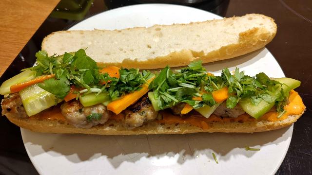 Biały talerz, na nim półbagietka ze sriracha mayo i pulpecikami wieprzowymi, marynowaną marchewką, ogórkiem i kolendrą