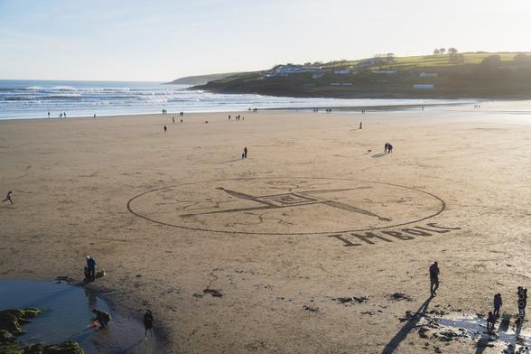 Beach landscape: A large St. Brigid's Cross design drawn in the sand with the text "IMBOLC" at Inchydoney Beach, with people walking along the shoreline, waves breaking in the distance, and houses visible on green hills in the background on a bright winter's day.