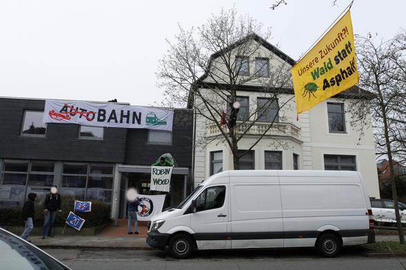 Großes Banner "Bahn statt Autobahn" an Gebäude und kleineres gelbes Banner "Unsere Zukunft - Wald statt Asphalt" über die Straße gespannt. 
Weitere kleine Banner von Robin Wood und KlimaKollektiv und Fahnen mit durchgestrichenen A39 und A20 Schildern.