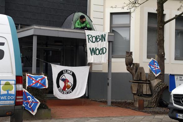 Aktivist von Robin Wood sitzt in Zelt auf Vordach der Autobahn GmbH und hält Robin Wood Banner. Darunter ist Banner vom KlimaKollektiv gespannt und kleine Fahnen mit durchgestrichenen A39 und A20 Schildern.