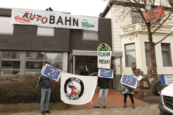 Großes Banner "Bahn statt Autobahn" an Gebäude der Autobahn GmbH. 1 Aktivisti sitzt auf Vordach übern Eingang mit Zelt und Robin Wood Banner, weitere Aktivisti halten kleine Fahnen mit durchgestrichenen A39 und A20 Schildern und KlimaKollektiv Banner und 1 Aktivisti ist im Baum mit "Refugees Welcome" Banner.