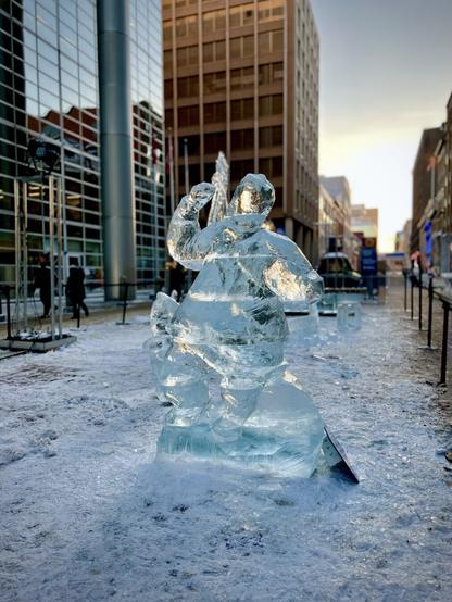 The image depicts a large ice sculpture situated in an urban environment, surrounded by snow and ice. The sculpture features a figure that appears to be a person, possibly in a dynamic pose, holding an object above their head. The ice is clear and reflects light, creating a shimmering effect. In the background, there are modern buildings with glass facades, indicating a bustling city atmosphere. The ground is covered in a mix of snow and ice, contributing to the wintery scene. The overall setting suggests a festive or artistic display, likely part of a winter event or festival.