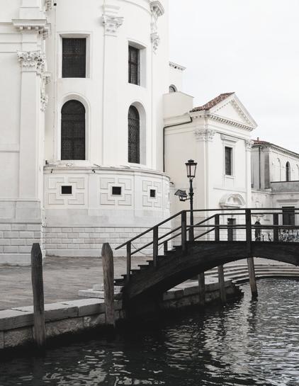 This photograph captures an architectural scene in Venice, Italy, blending elegance with timeless charm. The focal point is a beautifully ornate building with bright, white facades adorned with classical architectural details such as arched windows, decorative cornices, and intricate moldings. The structure exudes a sense of grandeur, characteristic of Venetian Renaissance and Baroque influences.

In the foreground, a quaint wooden bridge gracefully arches over a canal, its dark, weathered texture contrasting with the light tones of the building. Wooden mooring posts flank the edge of the water, grounding the scene in its Venetian context. The canal’s surface reflects the soft light of the overcast sky, adding depth and a sense of tranquility to the composition.

A wrought-iron lamp post near the bridge adds an additional touch of classic Venetian charm, while the muted palette of whites, greys, and browns gives the image a serene and harmonious aesthetic.