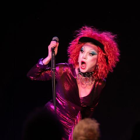 Felix La Freq in a sparkly purple/pink number, holding a microphone, with an expression of pure shock on their face
