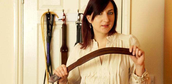 Pandora Blake with a serious expression, holding a leather strap and with a variety of other implements hanging behind them. Take your pick!