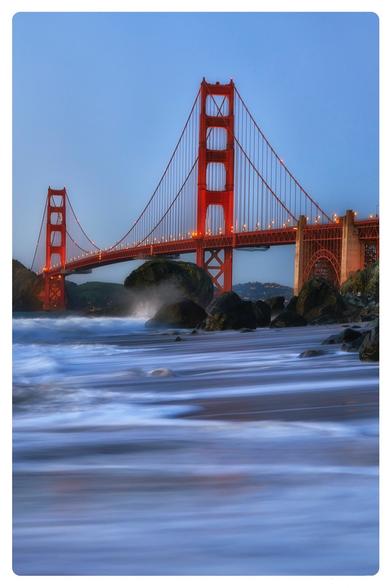 A scenic view of the Golden Gate Bridge at dusk, with the bridge's iconic red color standing out against a blue sky. Waves crash on rocky shorelines in the foreground, creating a tranquil atmosphere.