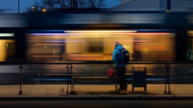 An evening scene at a bus stop. A person with a blue jacket and a red backpack is waiting at a bench. The person is photographed from behind while a tram rushes by. Due to the slow shutter speed the tram is very blur and more or less only visible as lines and stripes.