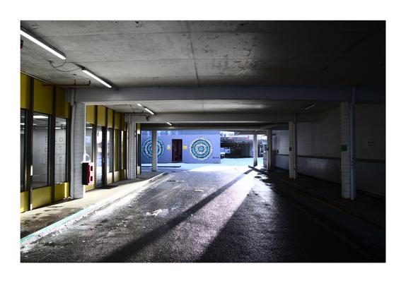 A dimly lit urban passageway with concrete pillars and yellow accents on the walls leads to an open courtyard. Sunlight streams through, creating a contrast of light and shadow. In the background, a mural with intricate circular patterns in shades of blue and white decorates the far wall.