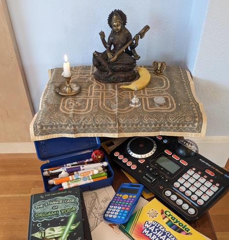 Saraswati idol surrounded with a kid's books, markers, and music/art supplies