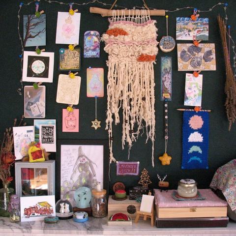Photo of altar during day time with postcards on the wall, woven wall art, crochet moon phases banner, a drawing of a witch, some tins and jars, and crafted watermelons for Palestine.
