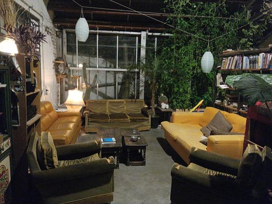 Plusieurs fauteuils très usés disposés en rond autour de plusieurs tablettes dans un bâtiment qui semble être un ancien garage. Plusieurs plantes vertes montent jusqu'au plafond et une bibliothèque de bandes dessinées sur la droite.