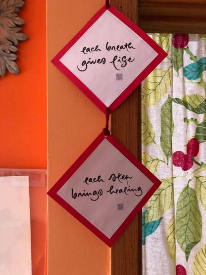 two squares hanging on red thread one above the other. Each square has white paper with red edging and black calligraphy. The top square read each breath gives life. The bottom square reads each step brings healing