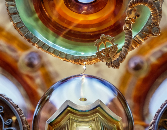 Close-up of a spark between a round glass-encased component to a slice of agate-like striped polished mineral fringed with tiny metal cubes, glowing orange and green with ornate details against a blurred background. Macro photography style.