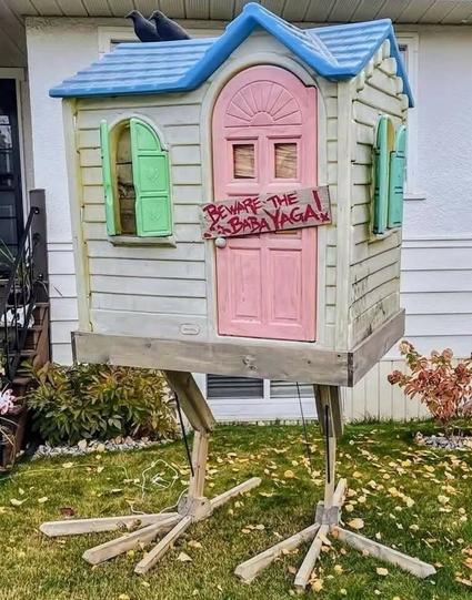 A plastic yard toy playhouse with  cute pastel accents has been altered: two ravens are on the roof and chicken feet (made of wood) hold the whole thing up. There's a wooden plaque on the door that says "Beware the Baba Yaga!" in all capitals & red ink