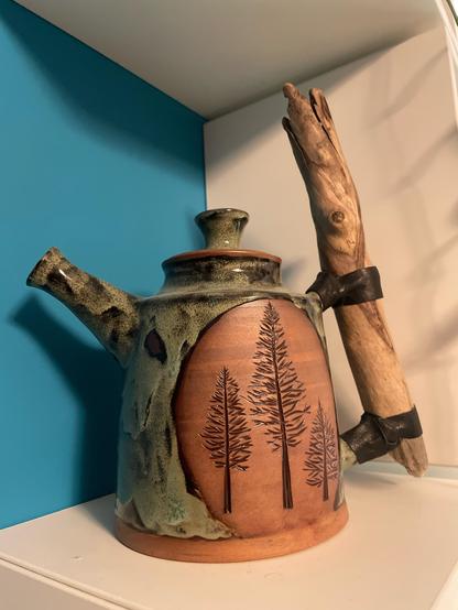 A red ceramic teapot. An emblem of exposed clay features black glaze in a carved forest scene. The rest of the teapot is a moody, lichen-y green. The handle is salvaged natural wood bound on with leather.
