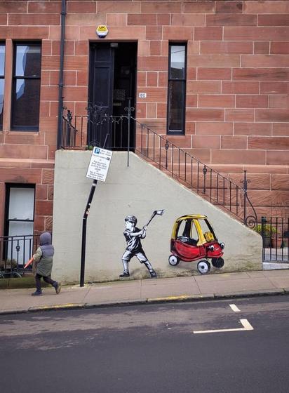 Streetart. A funny mural was sprayed with stencils on the concrete wall of a staircase in front of a street in Glasgow. There is also a traffic sign on the wall with a kink in the metal column showing parking times and payment information. Next to it is a mural of a little boy in black and white. He is wearing shorts and a T-shirt and is holding an axe. Behind him is a red and yellow children's car that looks like a golf cart with a number of fines on it. He is obviously very annoyed about the parking fees, because it looks as if he has hit the kink into the real traffic sign. (The photo also shows a child walking along the sidewalk)