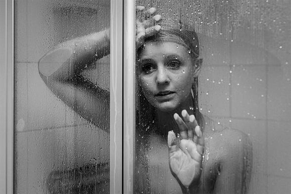 Junge Frau unter der Dusche, sie lehnt sich traurig, nachdenklich an die durchsichtige Duschwand, stützt die Hände dagegen und schaut in die Ferne.