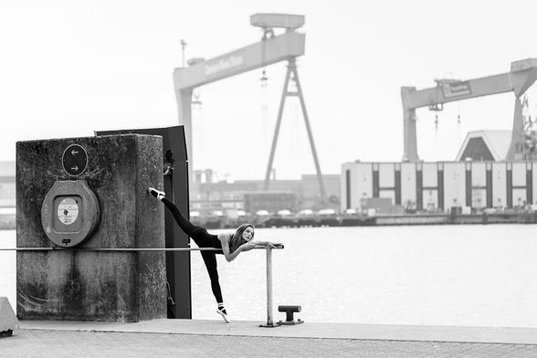 Balletttänzerin an der Hafenkante der Kieler Förde, im Hintergrund die Portalkräne der Werft. Sie steht auf einem Bein auf Spitze, das andere ist an einen großen Beton-Abstandshalter gelehnt, der die Schiffe von der Kaimauer abhalten soll.