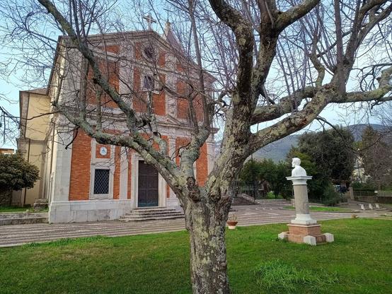 Facciata di una chiesa fotografata attraverso i rami di un albero reso spoglio dalla stagione invernale. In basso a destra, un busto marmoreo, poggiato su un piedistallo, è rivolto verso l'ingresso.