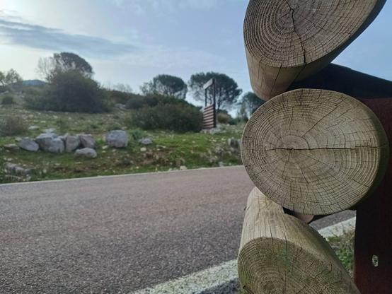 Tratto di strada asfaltata in buono stato. Sullo sfondo, fuori fuoco, la natura con alberi radi, rocce e manto erboso. In primo piano, a destra, la parte finale di un guardrail in legno e metallo.