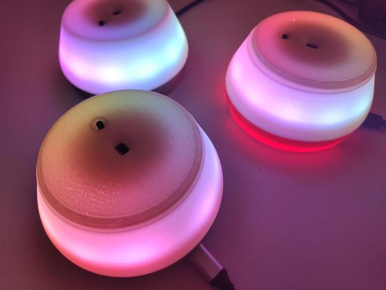 Nahaufnahme mehrerer leuchtender GlowLight-Nodes mit einem sanften Farbverlauf aus Lila, Blau, Rot und Gelb. Die Lampen haben ein modernes, rundes Design mit transluzenten 3D-gedruckten Gehäusen und leuchten in verschiedenen Farben. Ein angeschlossenes Kabel ist sichtbar, das die Stromversorgung der Geräte ermöglicht.