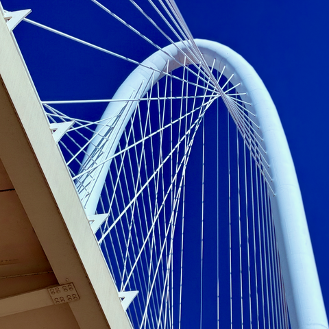 A close-up view of the Margaret Hunt Hill Bridge, seen from below, showing the weave of suspension cables hanging down from a large white metal loop, in a pattern which, from this angle, almost seems to spiral outward from the loop.