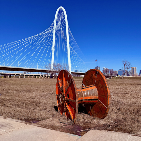 An empty orange metal spool roughly two meters in diameter, of the type which often holds cables, partially sunk into the soggy brown grasses of a river flood plain. Behind it can bee seen a modern white suspension bridge and part of a city skyline.