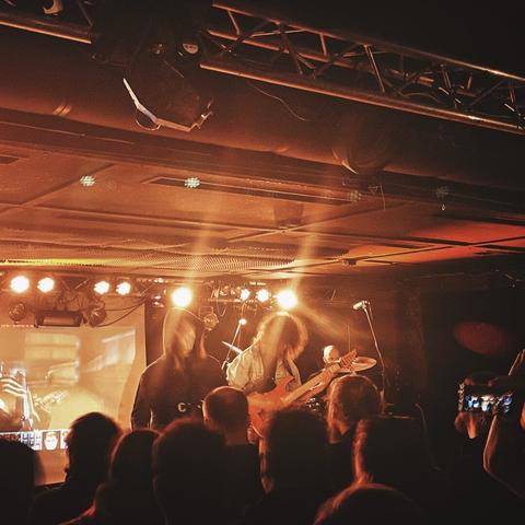 Gig photo. Two artists on the stage, in the middle of playing a song. One of them has a red guitar. Crowd watching.