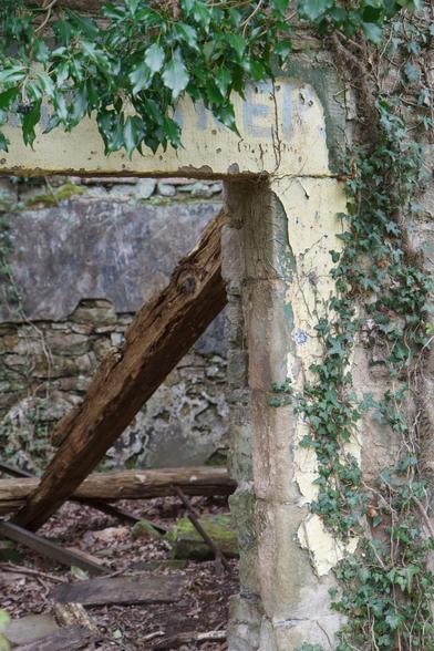 An old stone doorway, surrounded by ivy and partially covered in peeling paint - leading into a room open to the air, littered with debris and a fallen, rotting roof joist crossing the view at an angle. April 2014.
