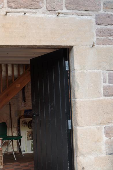 An old stone doorway, from the same angle as before - immaculately clean and surrounded by neat, rougher stonework - leading into a clean and tidy, enclosed room with a few items of furniture and the wooden structure of a staircase crossing the view at an angle. February 2025.