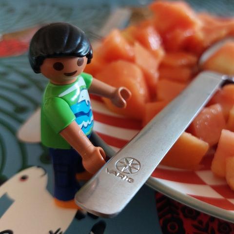 uma criança playmobil, de roupa verde e cabelo preto está junto de uma colher e um prato de mamão cortado aos pedaçoes